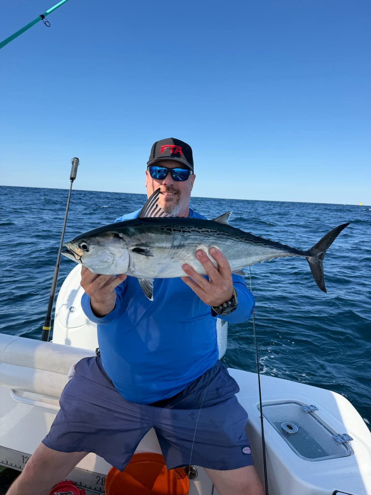 An angler in an FTA cap holding a bonito over the gunwale, deep blue offshore water behind.