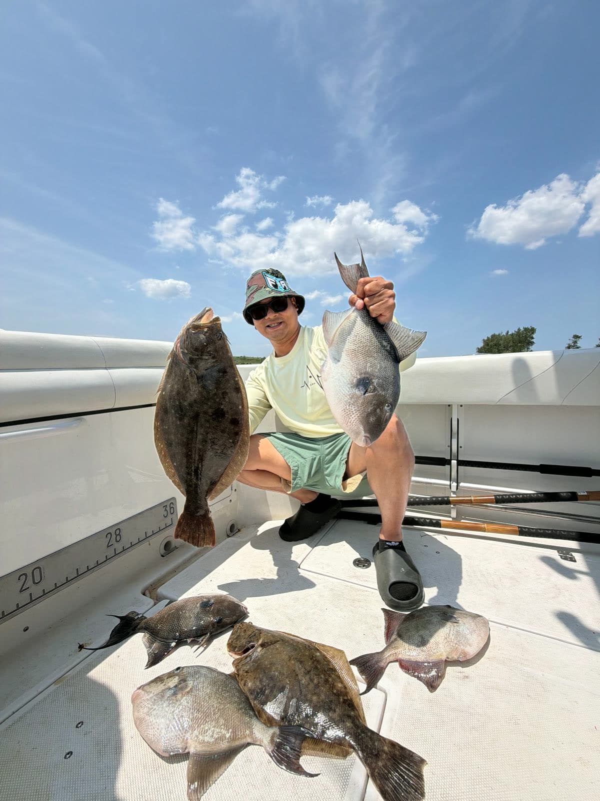 An angler crouched on the deck holding up a fluke and a sea bass, with more fish laid out across the deck and cumulus behind.