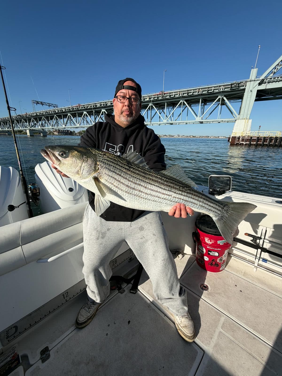 An angler holding a heavy striped bass at the rail with a steel bridge span crossing the water behind him.