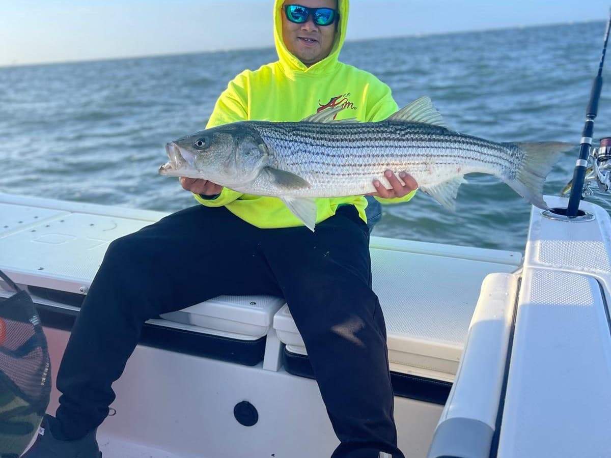 An FTA angler sitting on the gunwale holding a striped bass, open water behind.