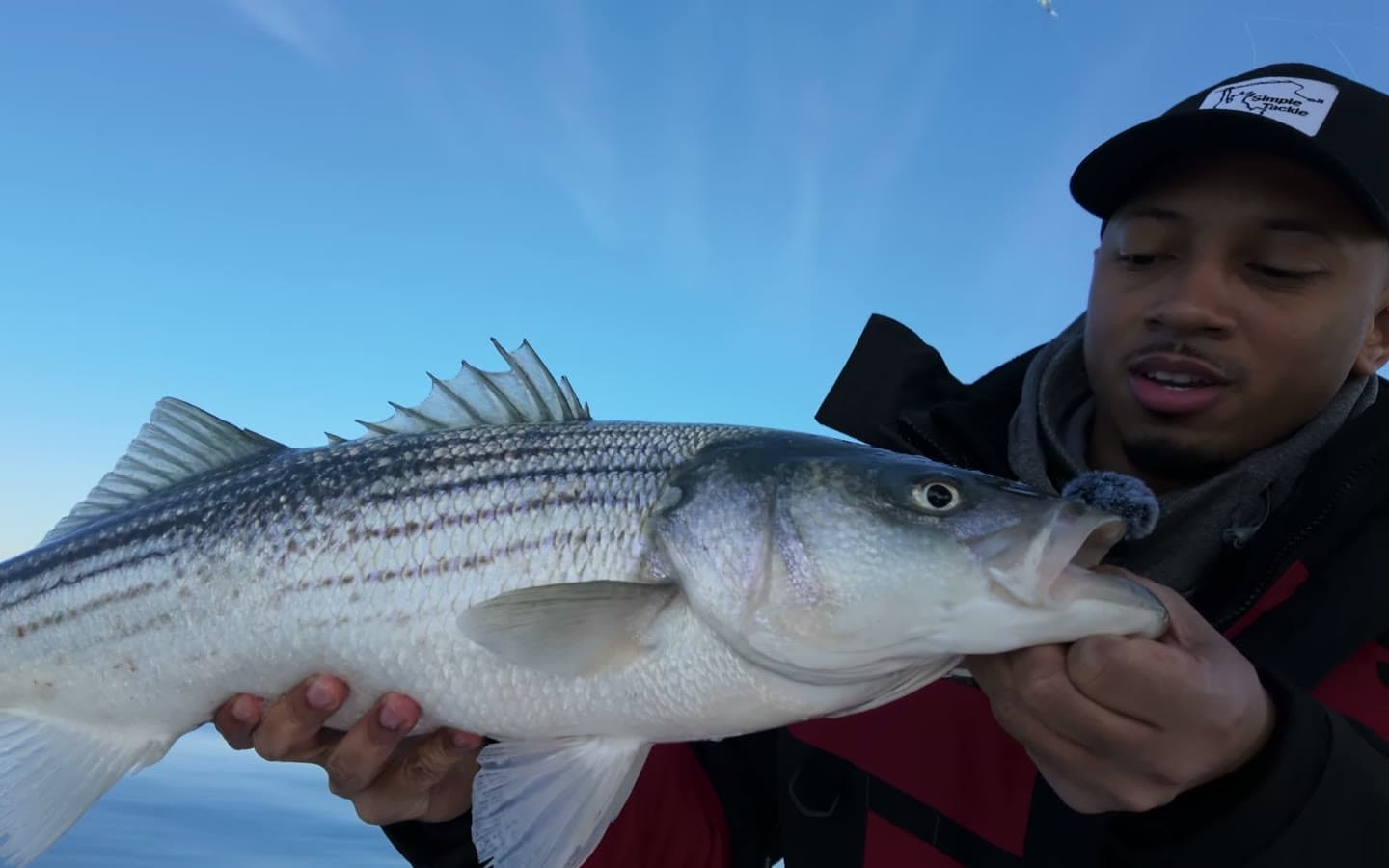 A striped bass held out across the frame, dorsal spines up, with the glassy bay and a clear winter sky behind.